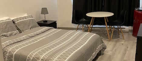 A bedroom with a bed, a nightstand, a lamp, a round table with two chairs, and a window with curtains.