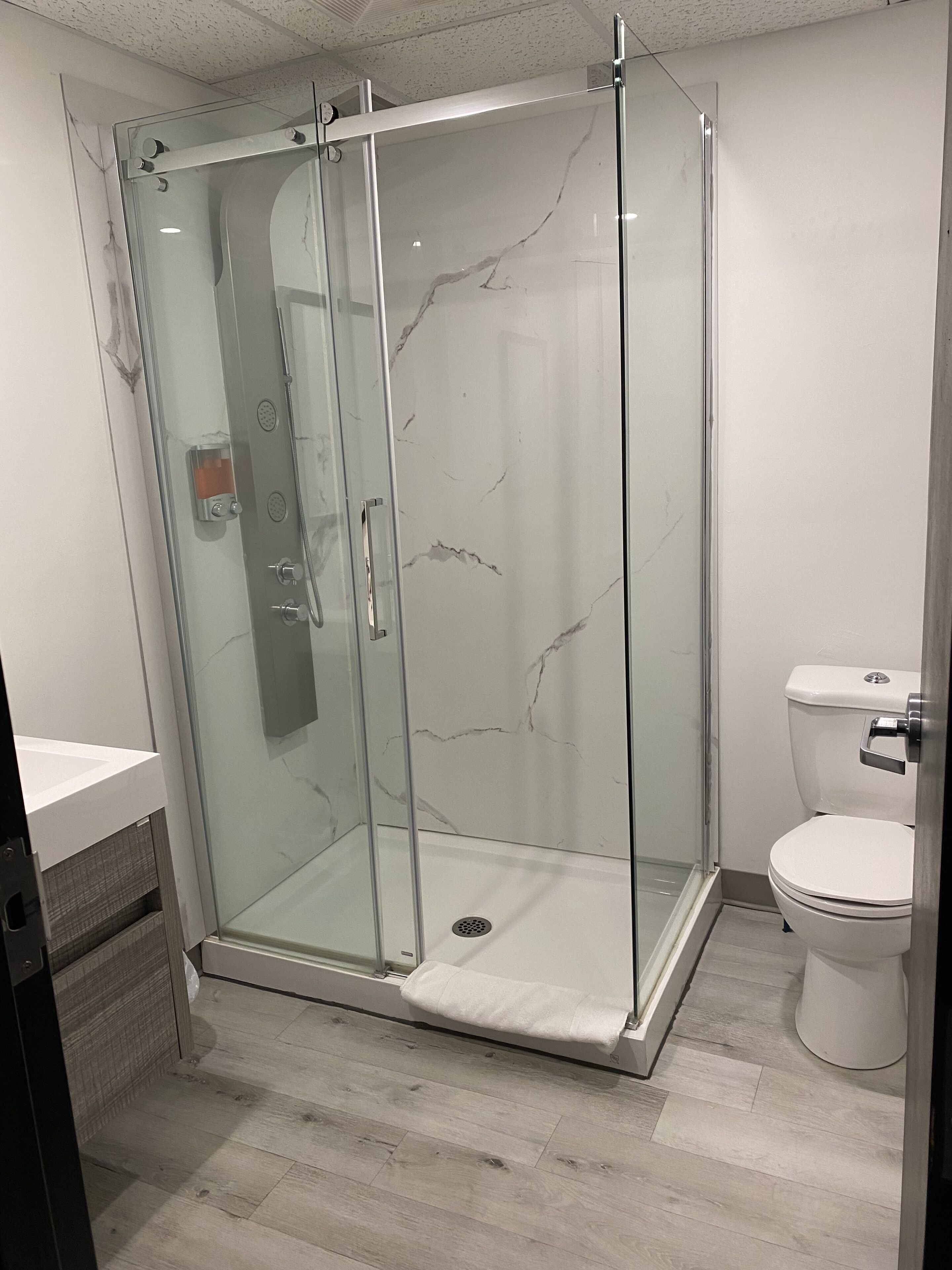A glass-enclosed shower with a marble wall, a white toilet, and a wooden floor.