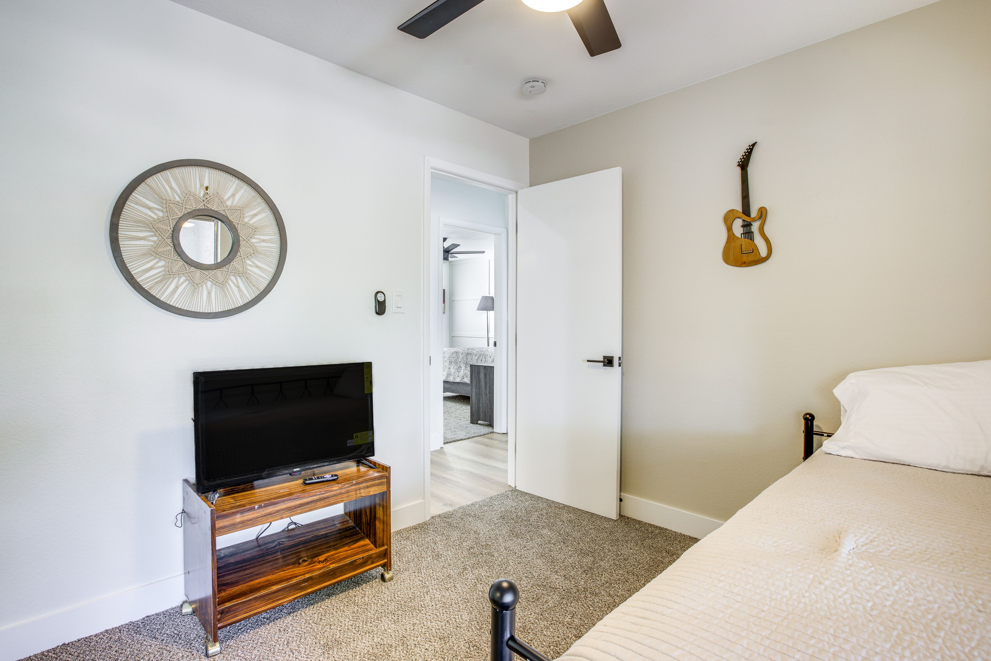 Un dormitorio con cama, televisor sobre un soporte de madera, guitarra colgada en la pared y una puerta que da a otra habitación.