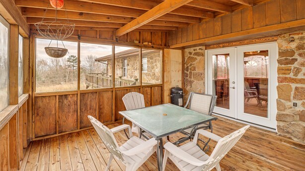 Una terraza de madera con una mesa y sillas, muros de piedra y ventanas amplias.