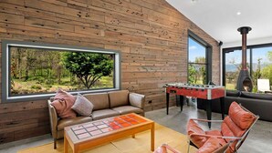 Ein gemütliches Wohnzimmer mit einer Holzwand, einem großen Fenster mit Blick auf einen Wald, einem Ledersofa, einem Kicker und einem Kamin.