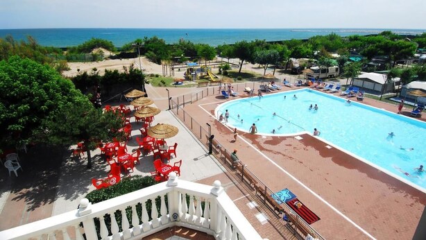 Ein Poolbereich mit Meerblick, umgeben von roten Stühlen und Sonnenschirmen, sowie ein Strand mit badenden Personen.