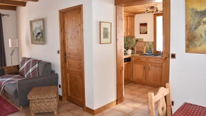 Una sala de estar acogedora con un sofá, un reposapiés y una puerta de madera que da a una cocina con armarios de madera y piso de baldosas.