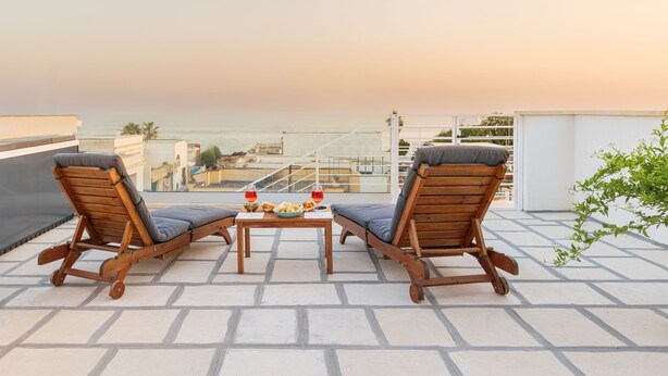 A terrace with two wooden lounge chairs and a table with a bowl of fruit and two glasses of red wine.