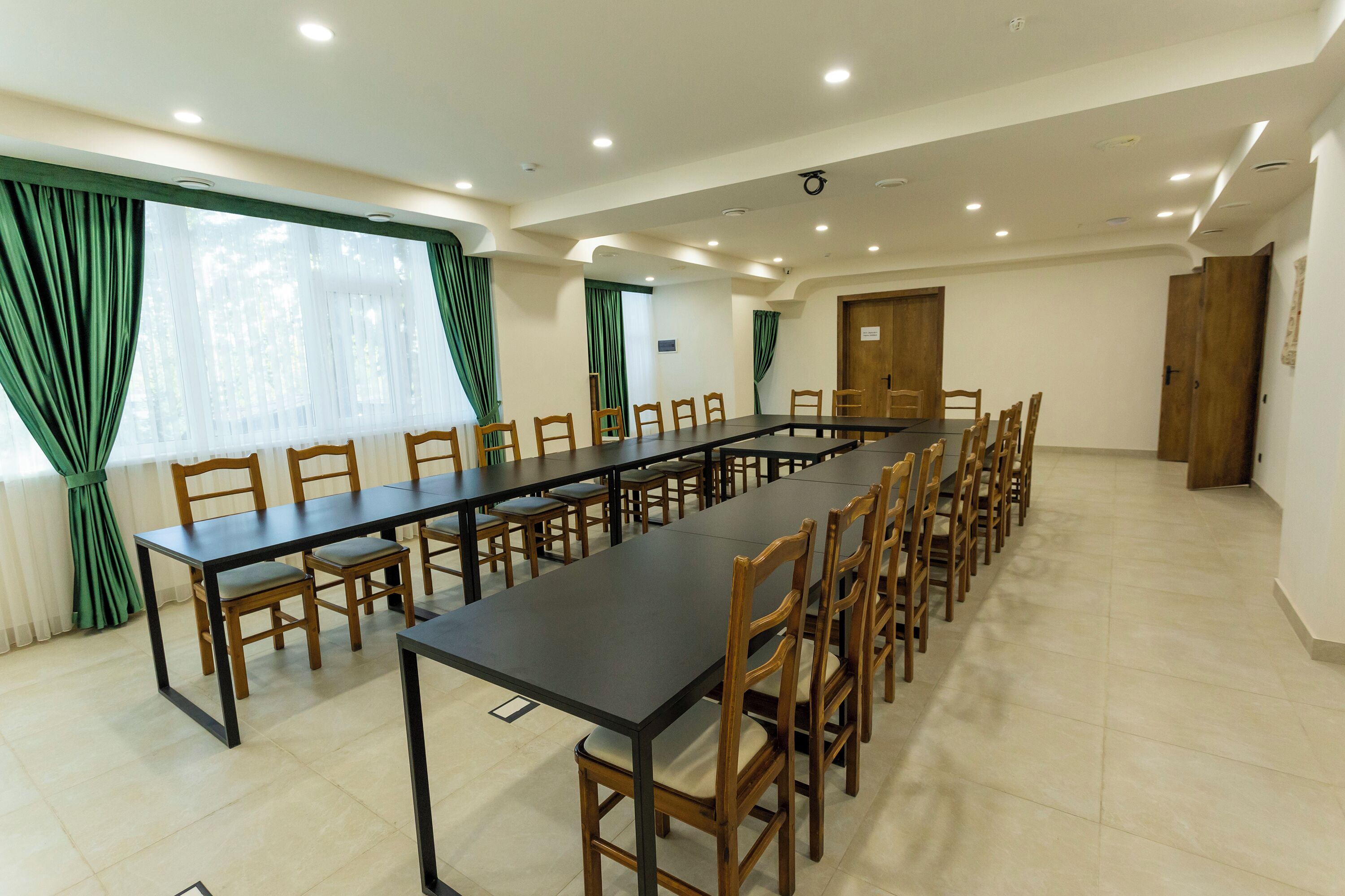 A conference room with a long table and multiple chairs, large windows with green curtains, and a tiled floor.