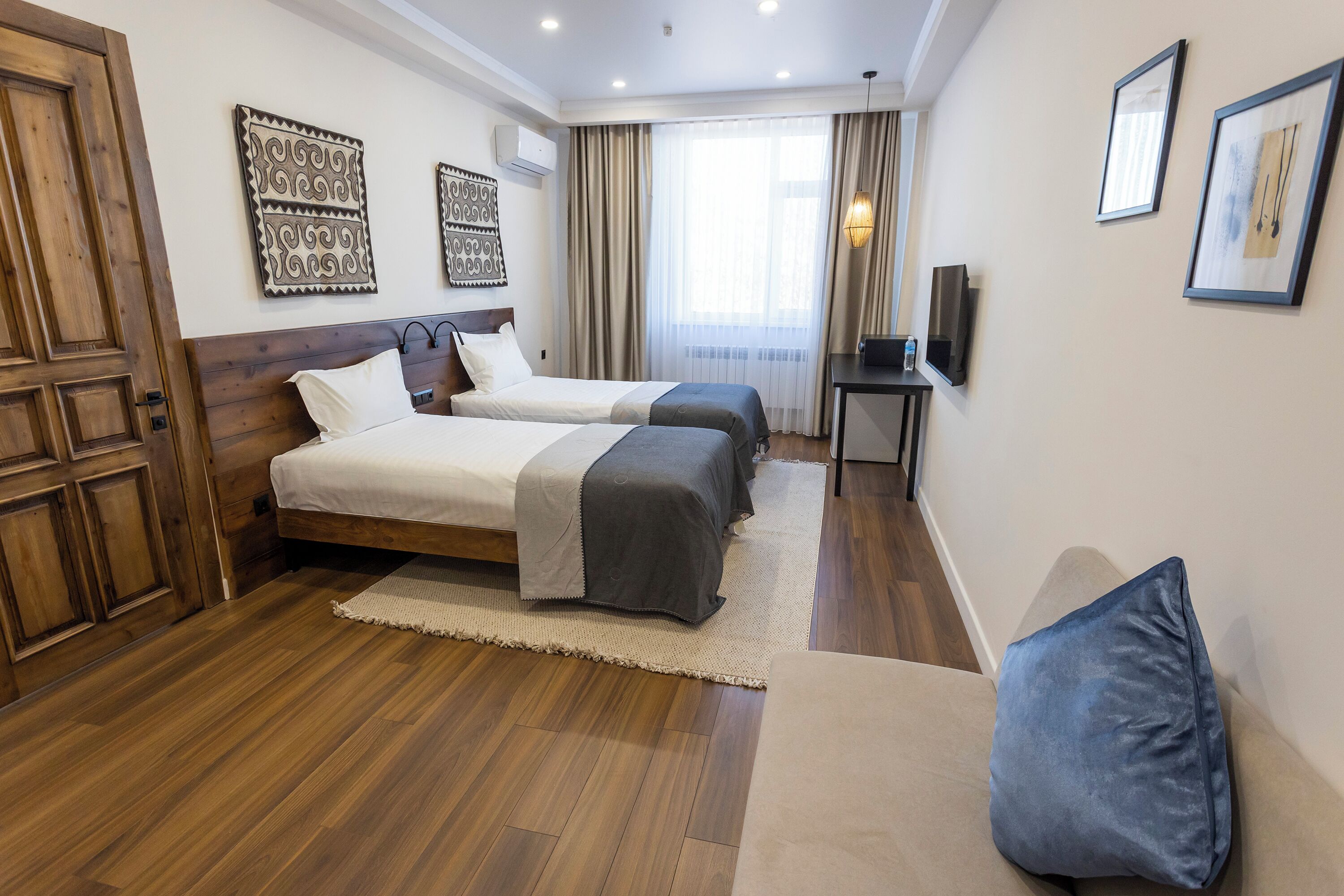Habitación de hotel con dos camas, piso de madera, un ventanal grande con cortinas y cuadros colgados en la pared.