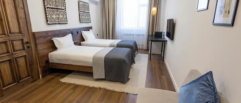 Habitación de hotel con dos camas, piso de madera, un ventanal grande con cortinas y cuadros colgados en la pared.