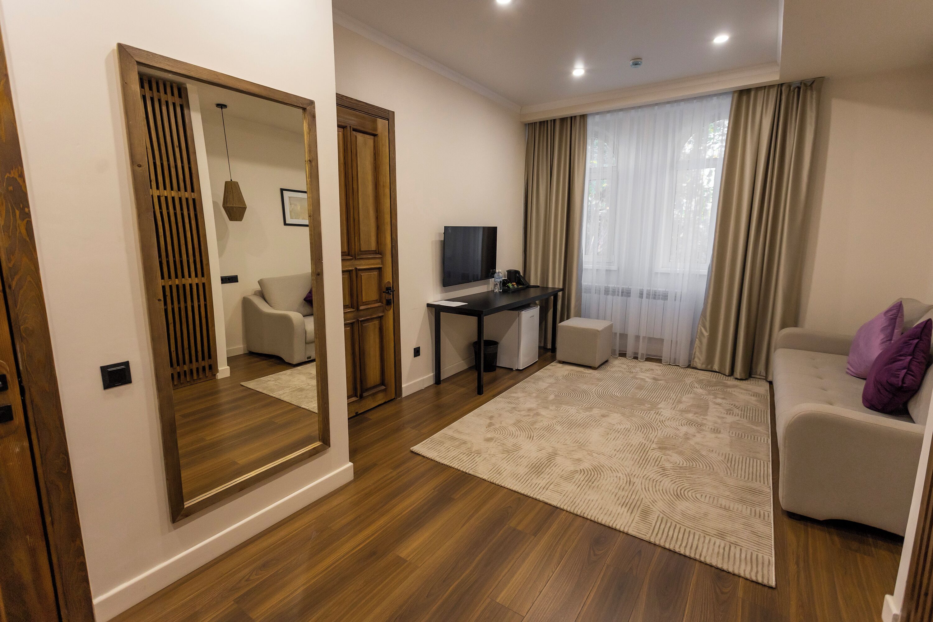 Una sala de estar moderna con piso de madera, un espejo grande, un sofá, un escritorio con televisor y un ventanal con cortinas.