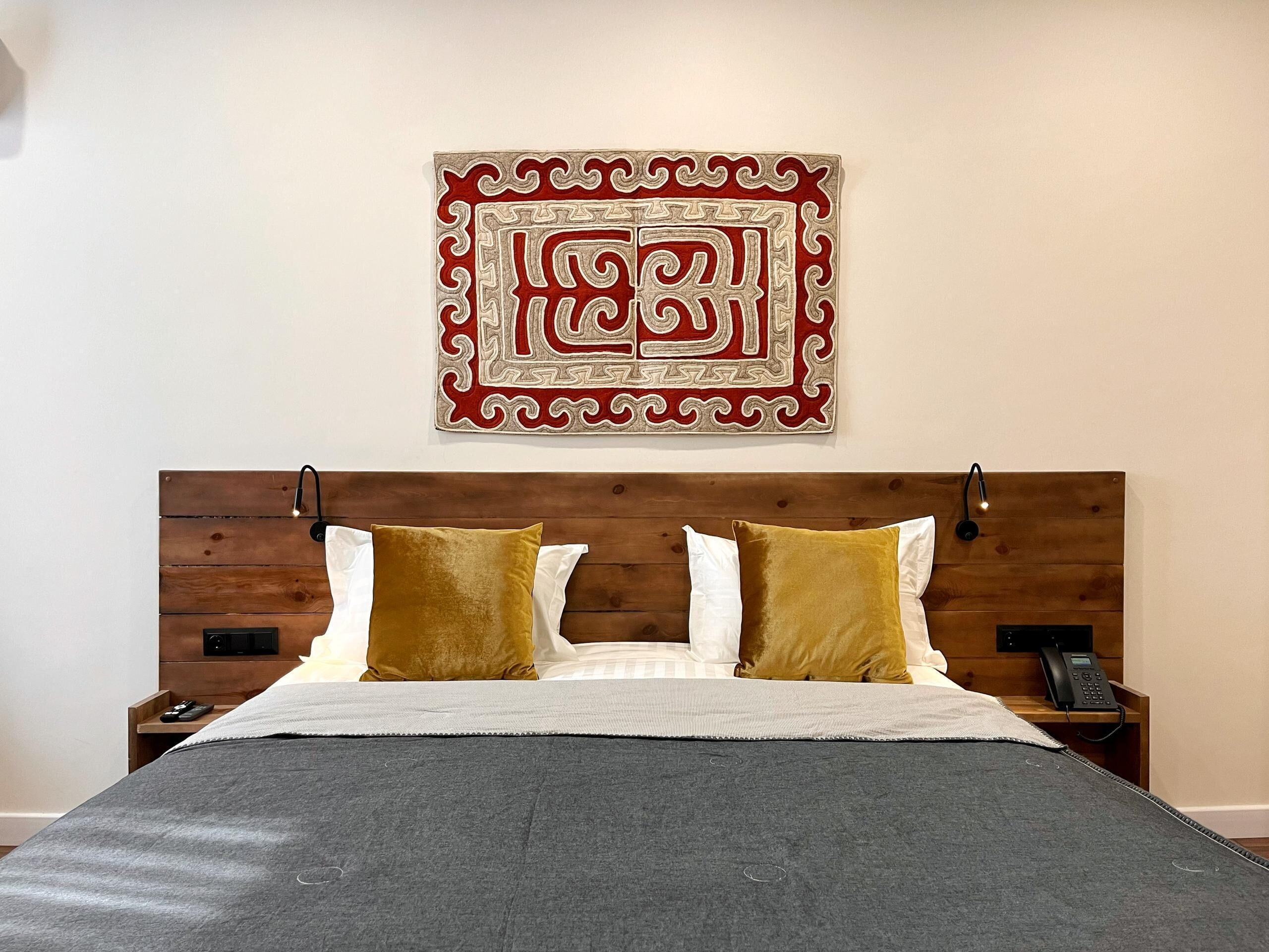 A neatly made bed with a grey blanket, two white pillows, and two mustard-colored pillows. A decorative wall hanging with a red and beige design is above the bed.