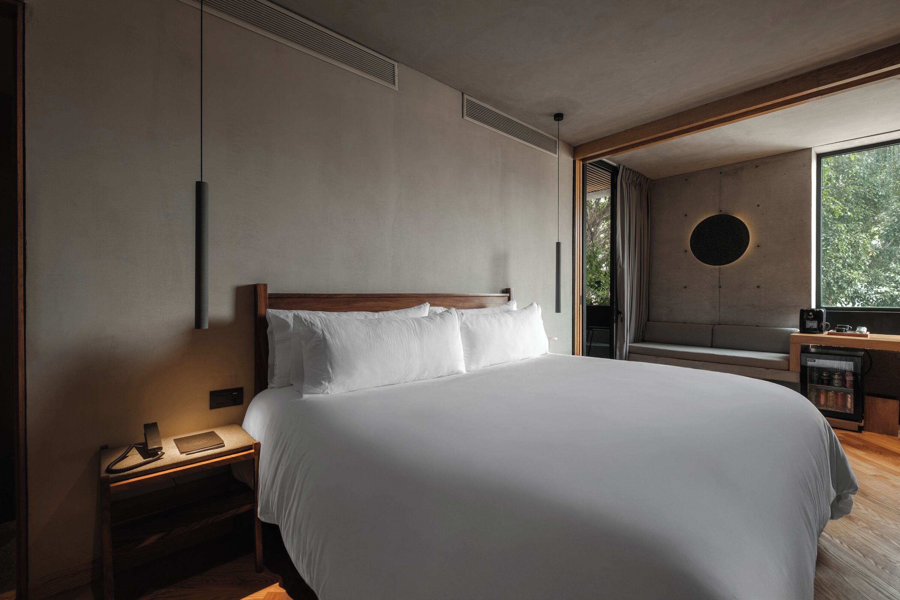 A modern bedroom with a large bed, a bedside table with a lamp, and a view of the outdoors through a window.