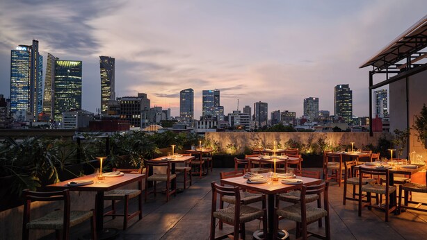 Un área de comedor al aire libre con varias mesas y sillas, con el perfil de un horizonte urbano al anochecer.