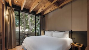 Ein Schlafzimmer mit einem großen Bett, einem Nachttisch, einer Lampe und Blick auf Bäume durch ein Fenster.