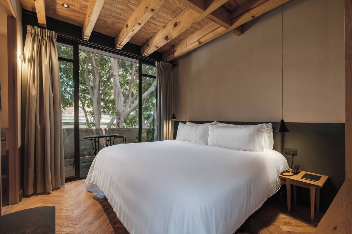A bedroom with a large bed, a nightstand, a lamp, and a view of trees through a window.
