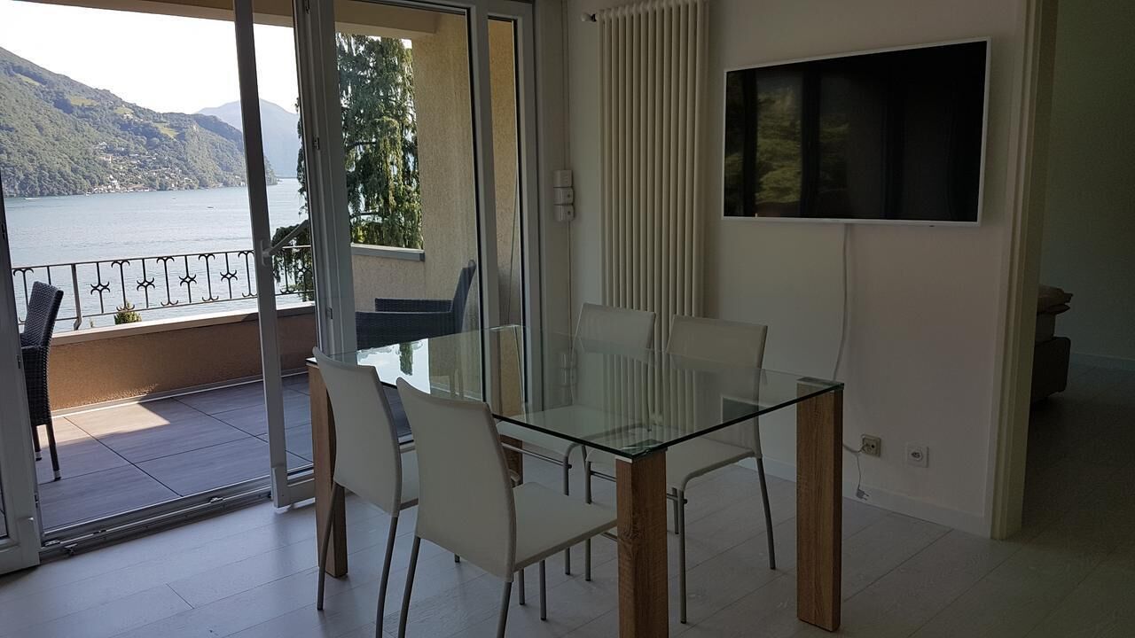 A modern living room with a glass table and chairs, a TV mounted on the wall, and a view of a lake and mountains through a sliding glass door.