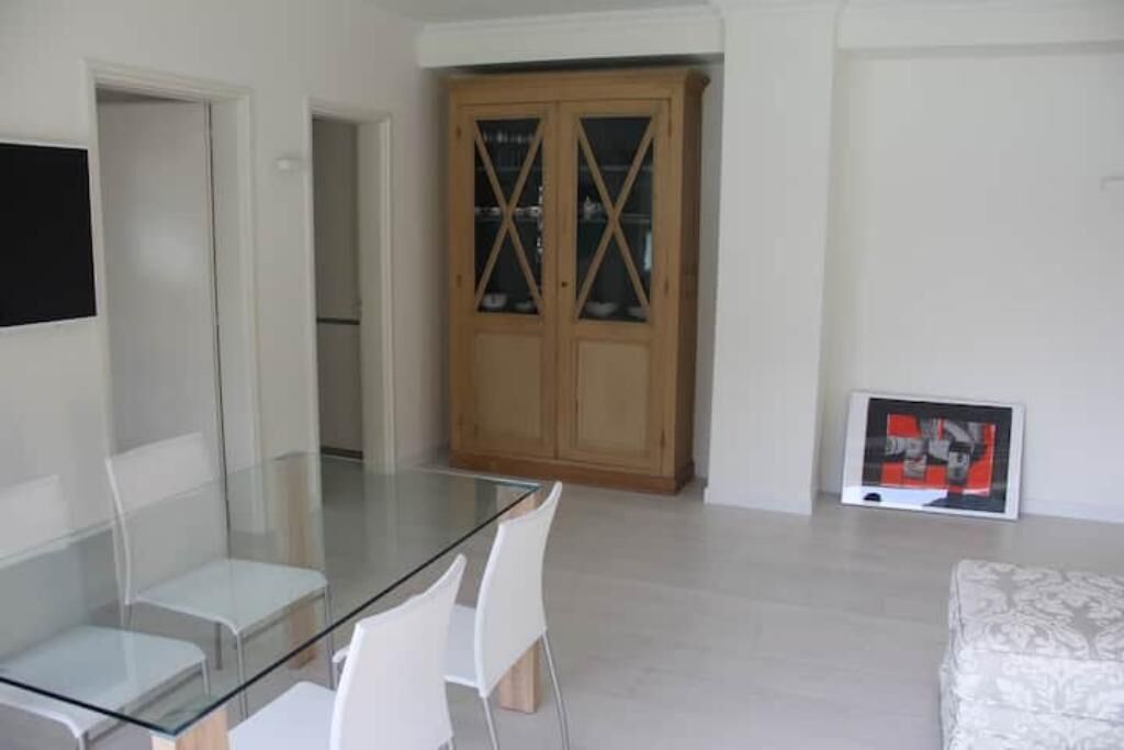 A modern living room with a glass dining table, white chairs, a wooden cabinet, and a framed picture on the wall.