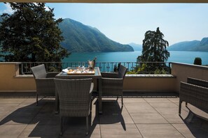 Eine Terrasse mit Korbmöbeln und einem Esstisch, mit Blick auf einen See und Berge.