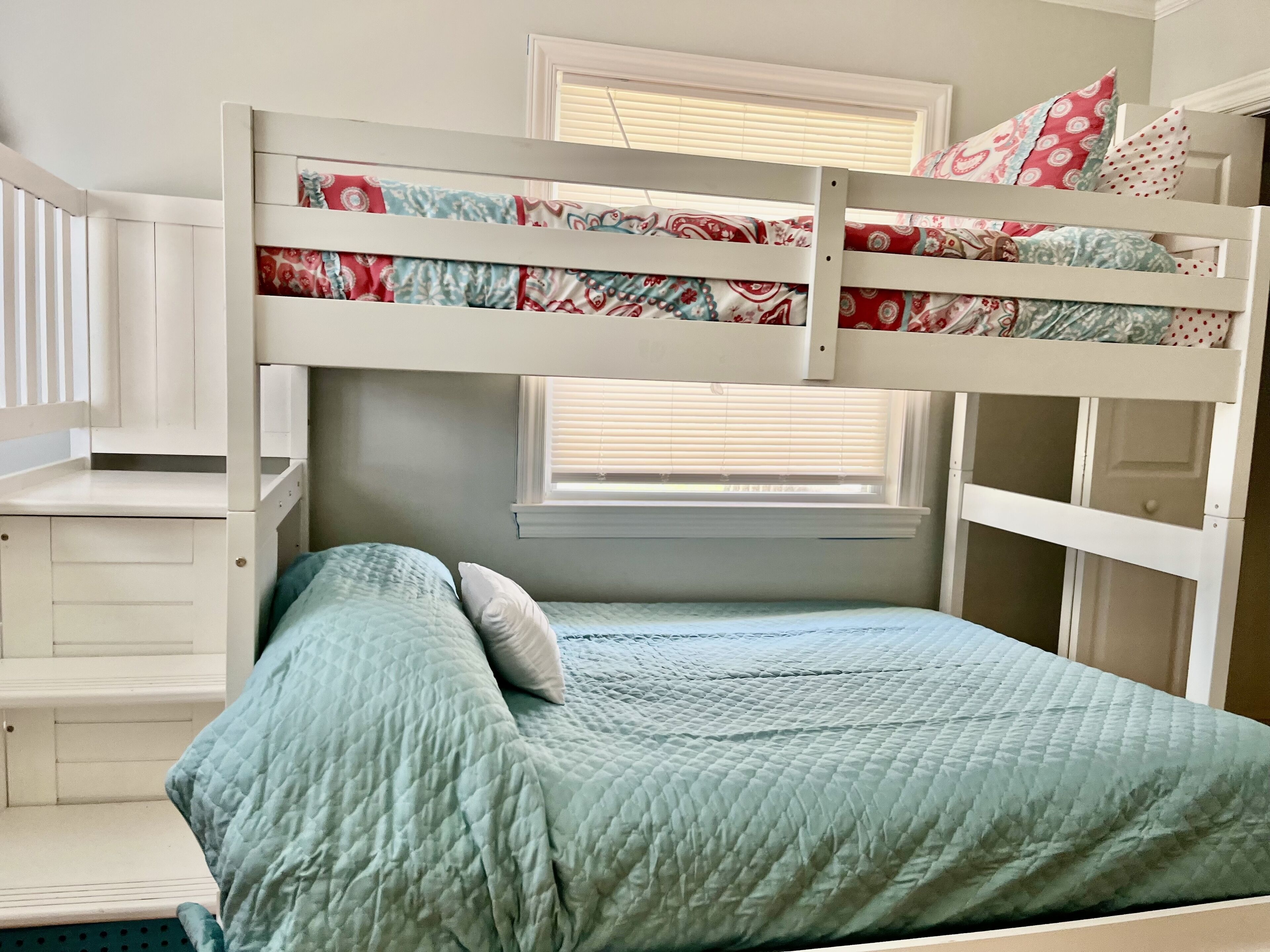 A bunk bed with a lower bed and a higher bunk, both with patterned bedding. There is a window with blinds, a white dresser, and a small shelf.