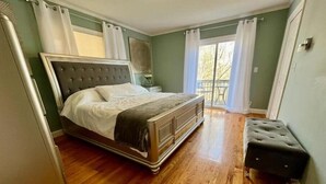 A bedroom with a large bed, a nightstand, a bench, and a view of the outdoors through a window.