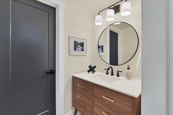 A bathroom with a dark door, a white vanity with a round mirror, and a light fixture above the mirror. - Pick of the Pines Pinehurst (Pinehurst)