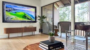 A modern living room with a large flat-screen TV, a wooden coffee table with books, a potted plant, and a view of the outdoors through a glass door. - Pick of the Pines Pinehurst (Pinehurst)