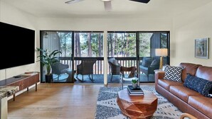 A spacious living room with a ceiling fan, a large flat-screen TV, a leather sofa, and a coffee table. There is a balcony with outdoor seating and a view of trees. - Pick of the Pines Pinehurst (Pinehurst)