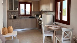 A compact kitchen with wooden cabinets, a microwave, a refrigerator, and a dining area with a table and chairs. - Villa Ersa @home by the sea (Santorini)