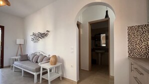 A modern living room with a sofa, a small table, and a decorative wall piece. An archway leads to a bathroom. - Villa Ersa @home by the sea (Santorini)