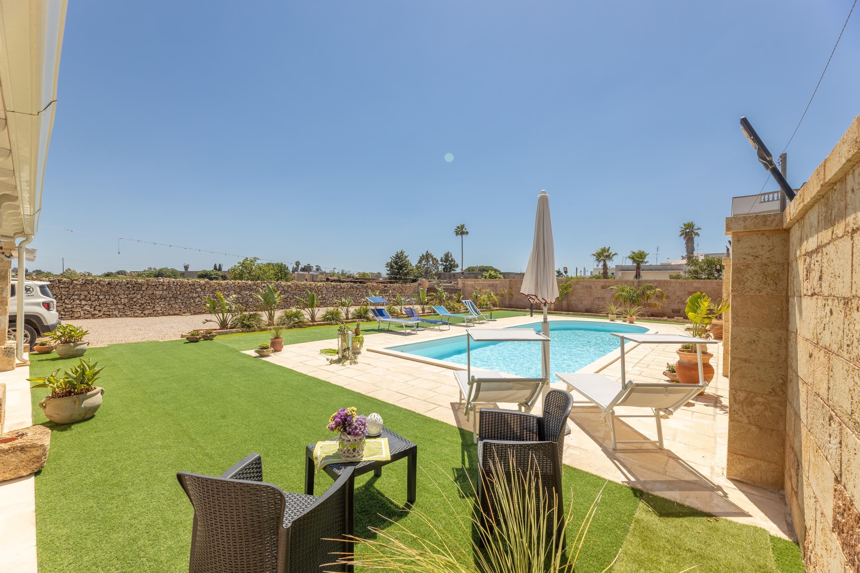 Un jardin avec une piscine, du gazon synthétique, du mobilier d’extérieur et un mur en pierre.
