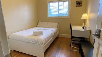 A small, clean room with a single bed, a desk, and a chair. There is a window with blinds, a lamp on the desk, and a bear silhouette on the wall.