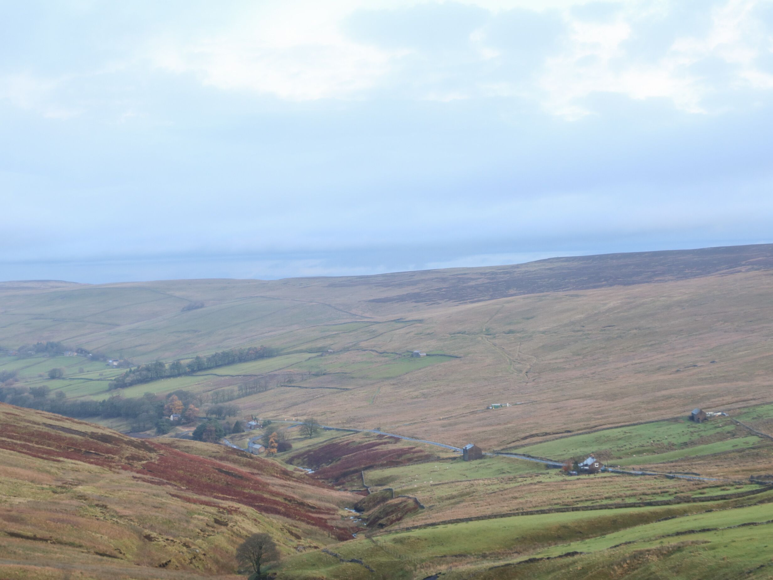 A panoramic view of a rural landscape with rolling hills, scattered houses, and a distant body of water under a cloudy sky.