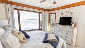 Een slaapkamer met een groot raam, een bed met witte lakens, een dressoir en een flatscreen televisie.