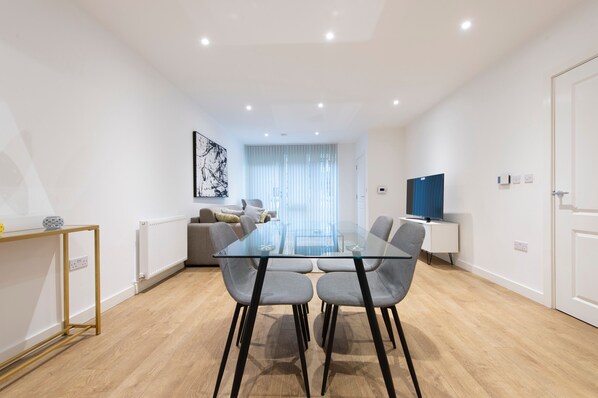 Un salon moderne avec une table à manger en verre, des chaises grises, un canapé, un téléviseur et une grande fenêtre ornée de rideaux légers.