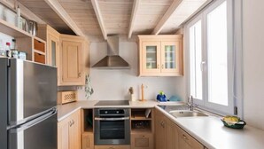 A compact kitchen with wooden cabinets, a stainless steel refrigerator, a built-in oven, and a window providing natural light. - House with sea view 'Avgi' @seaside house (Santorini)