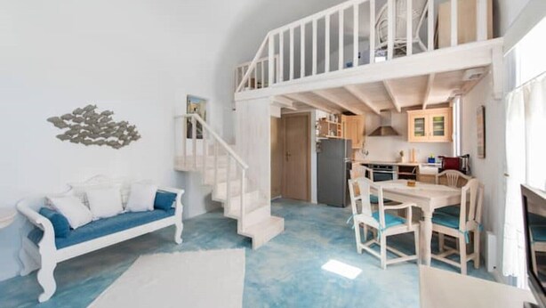 A spacious living area with a white staircase leading to a loft, a dining area with wooden furniture, and a kitchen with wooden cabinets. - House with sea view 'Avgi' @seaside house (Santorini)