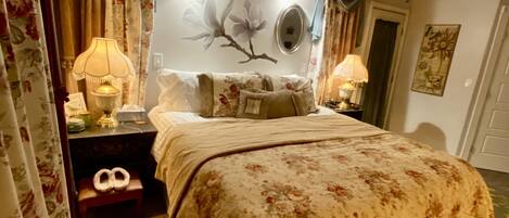A bedroom with a bed, two bedside tables, a ceiling fan, a rug, and a wall with a mirror and a floral painting.