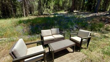 Een houten zitgedeelte buiten met een bank en twee stoelen, omgeven door een bebost gebied met hoge bomen en een grasveld met wilde bloemen.