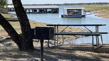 A picnic table and a trash bin near a body of water with a boat dock and buildings in the background.