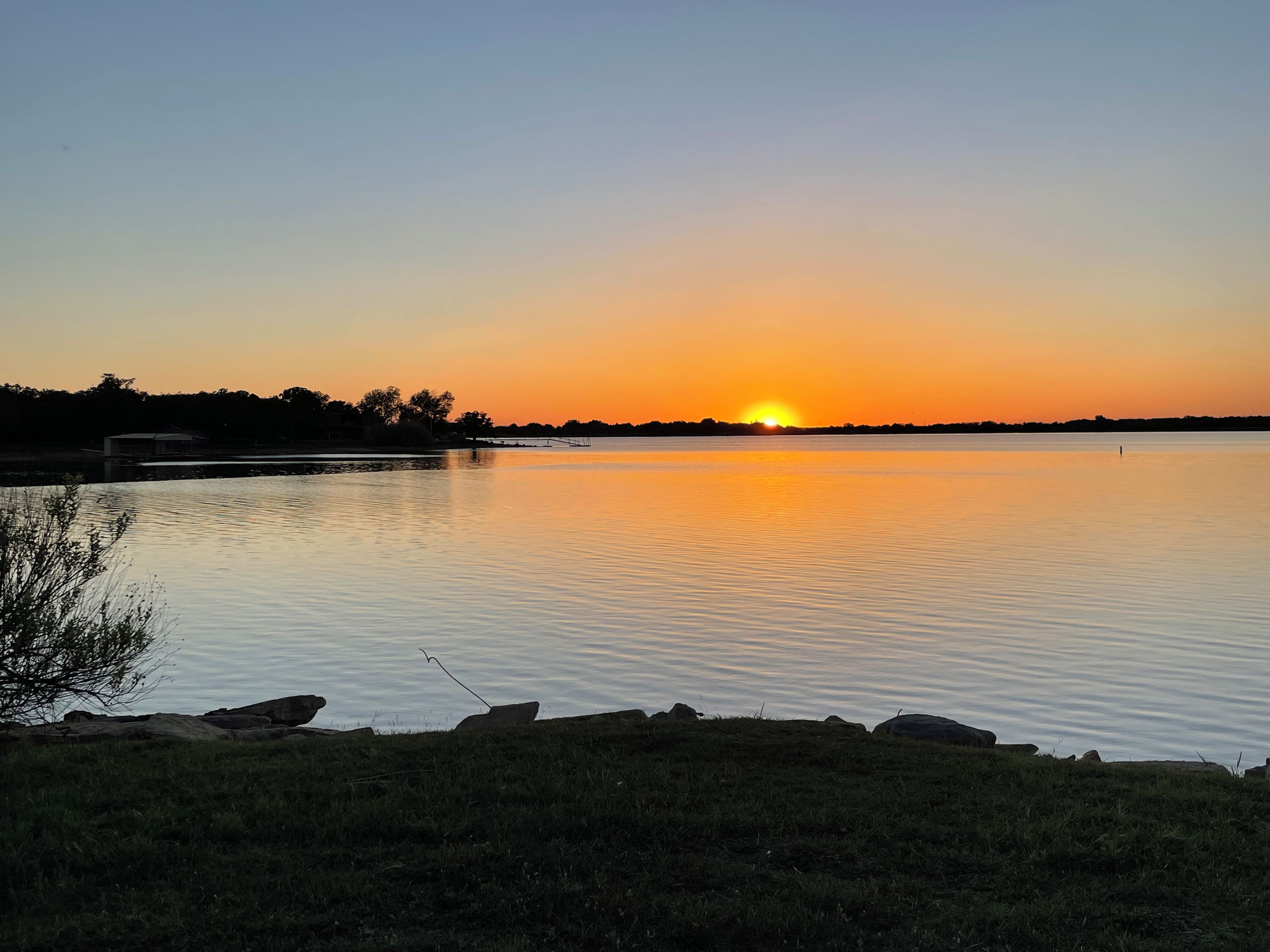 Ein friedlicher Sonnenuntergang über einem stillen See mit einer grasbewachsenen Uferlinie und Felsen.