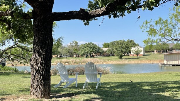 A serene outdoor area with a pond, two white Adirondack chairs, and a large tree providing shade.