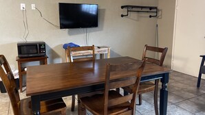 A dining area with a wooden table and chairs, a television mounted on the wall, a microwave on a small table, and a broom leaning against the wall.