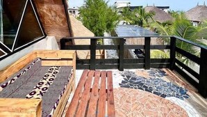 A wooden deck with a patterned rug and a bench, overlooking a garden with palm trees and a building with a thatched roof. - Lekki Beach Resort- 1 bed & private pool (Lekki)