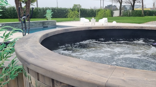A hot tub with a wooden edge, surrounded by a landscaped garden with benches and a pool.