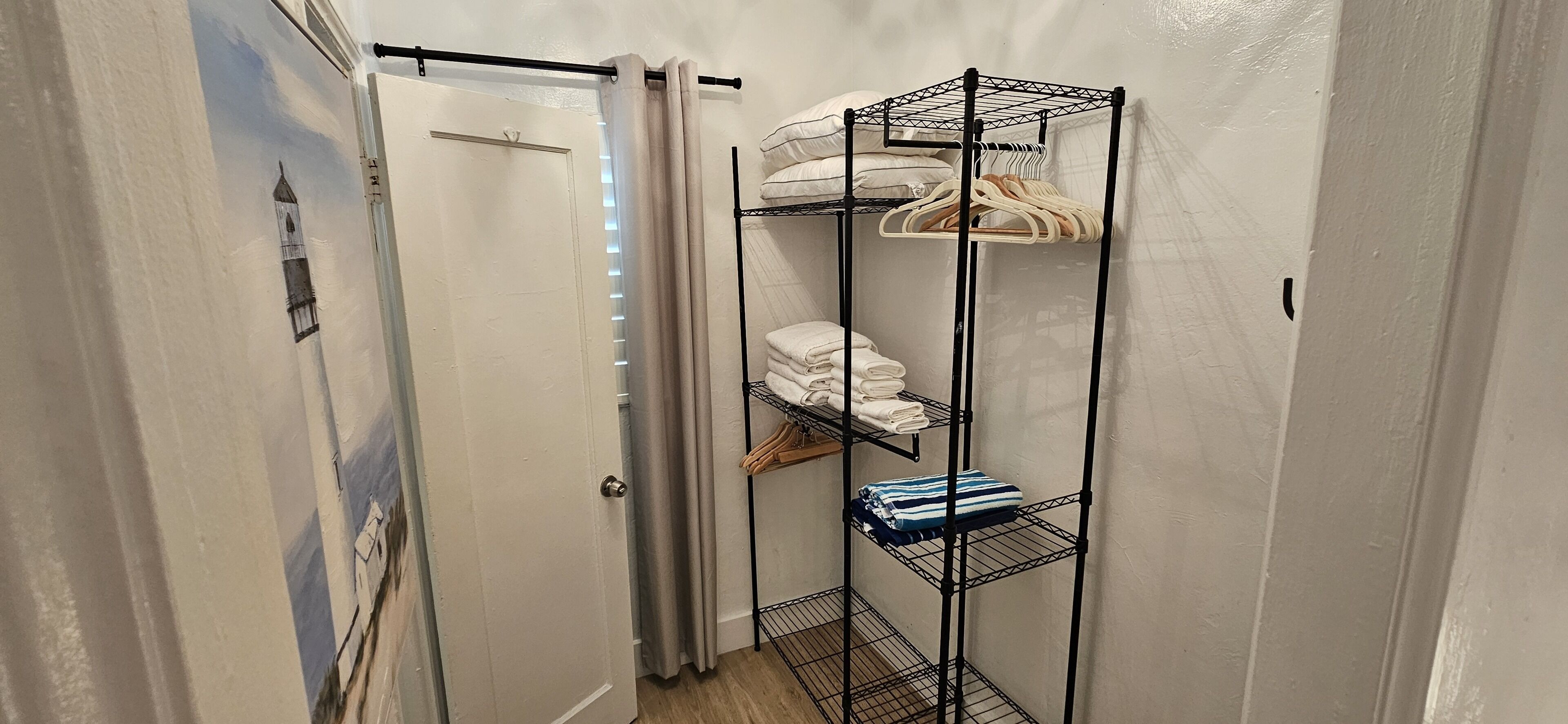 A narrow hallway with a door, a metal shelf unit with towels and hangers, and a mural on the wall.