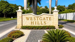 Ein Steinschild mit goldener Schrift 'WESTGATE HILLS', umgeben von Grünpflanzen und Palmen.