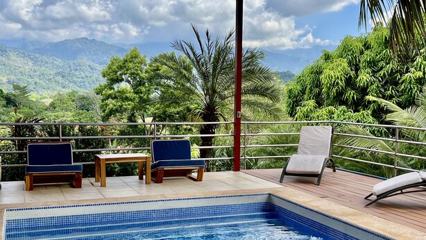 Uma área de piscina com duas cadeiras e uma mesa, rodeada por um deck de madeira e guarda-corpo de metal, com vista para uma paisagem exuberante com árvores e montanhas.
