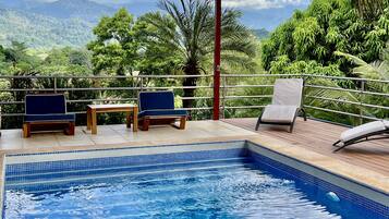 Un espace piscine agrémenté de deux chaises et d’une table, entouré d’une terrasse en bois et d’une rambarde en métal, offrant une vue sur un paysage luxuriant, avec des arbres et des montagnes.