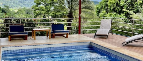 Un espace piscine agrémenté de deux chaises et d’une table, entouré d’une terrasse en bois et d’une rambarde en métal, offrant une vue sur un paysage luxuriant, avec des arbres et des montagnes.