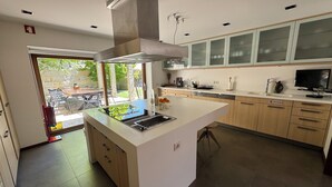 A modern kitchen with a central island, built-in cabinets, and a large window overlooking a patio area. - Quinta Trindade (Arouca)