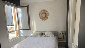 Un dormitorio con un ventanal grande, una cama con colcha a cuadros, un espejo montado en la pared y una mesita de noche con lámpara.