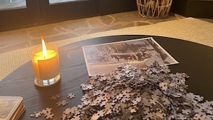Ein Zimmer mit großem Fenster, einer brennenden Kerze, einem Puzzle auf dem Boden und einem Holzkorb.